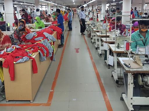 Ein Blick in eine Textilfabrik in Bangladesch. Ein Gang befindet sich zwischen einer Reihe von Tischen mit Nähmaschinen an denen Frauen sitzen und Kleidung herstellen.