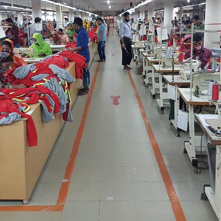 Ein Blick in eine Textilfabrik in Bangladesch. Ein Gang befindet sich zwischen einer Reihe von Tischen mit Nähmaschinen an denen Frauen sitzen und Kleidung herstellen. 