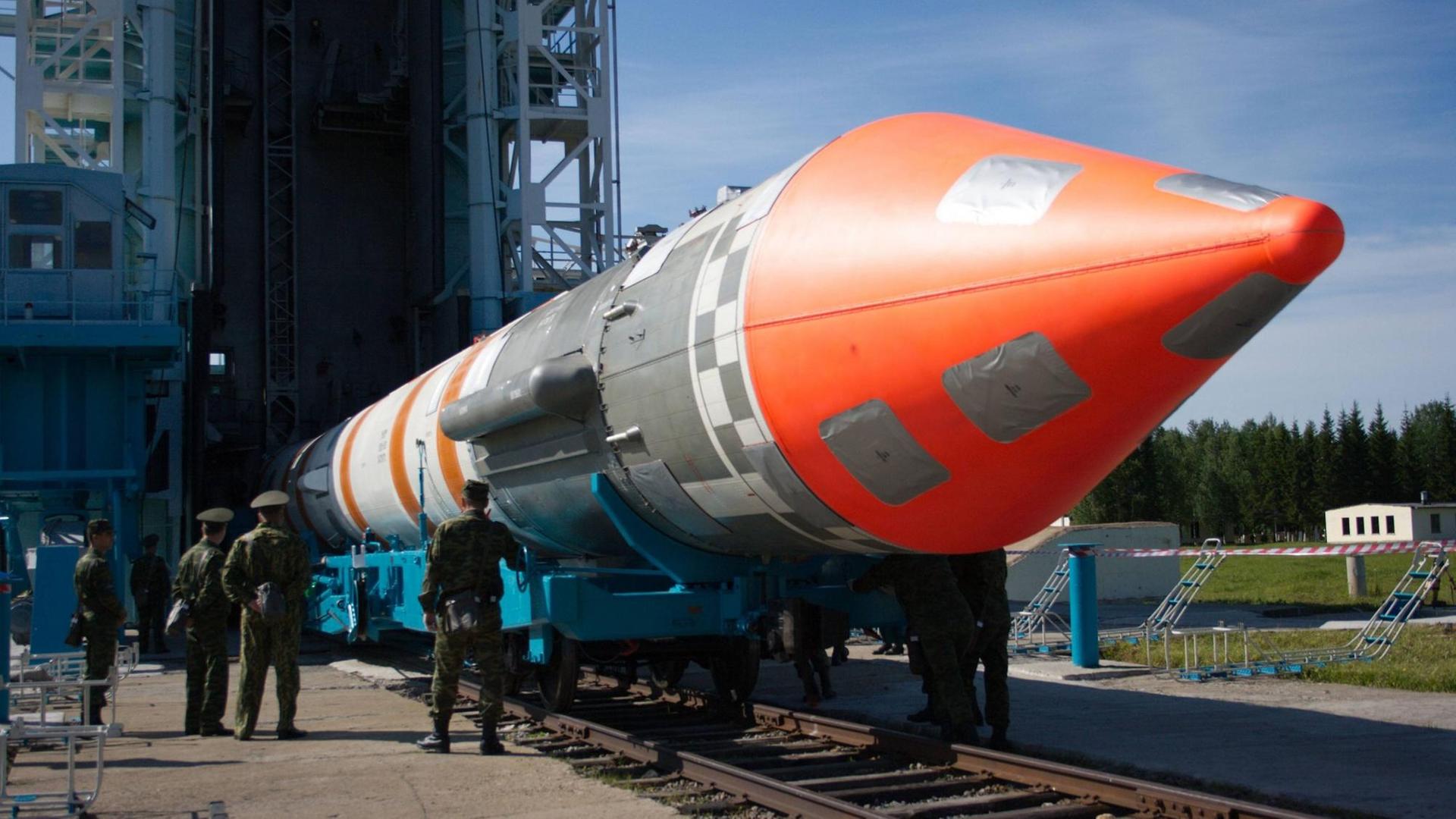 Eine riesige, rot-silberne Cosmos-3 Rakete liegt auf dem Boden vor einer Abschussrampe. Davor stehen drei russische Soldaten in Uniform.