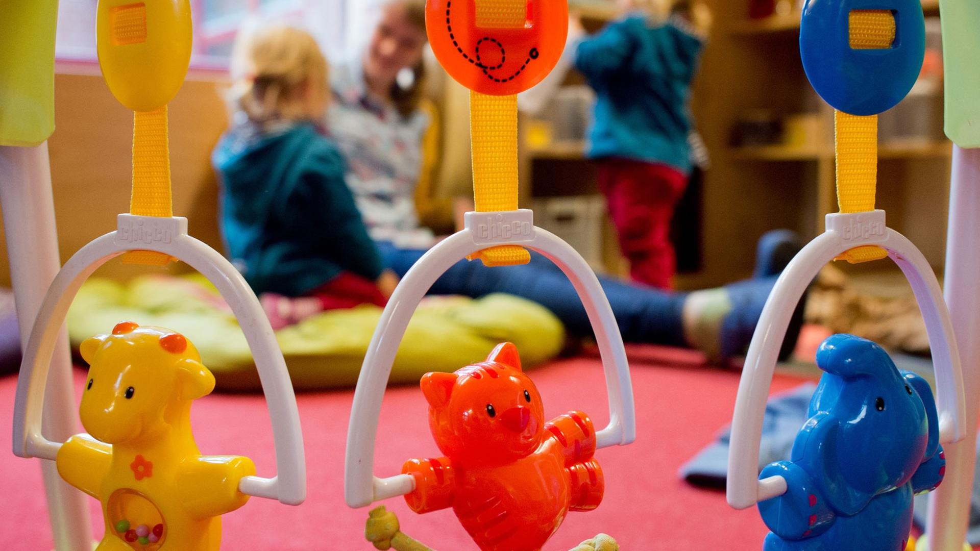 Kinderspielzeug hängt an einem Rahmen. Im Hintergrund spielt eine Erzieherin mit zwei kleinen Kindern