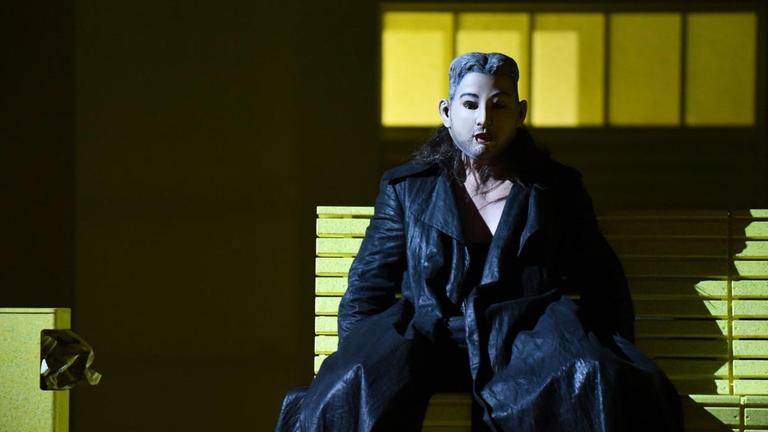Don Giovanni sitzt auf einer Parkbank, trägt eine Maske und schaut ins Publikum.