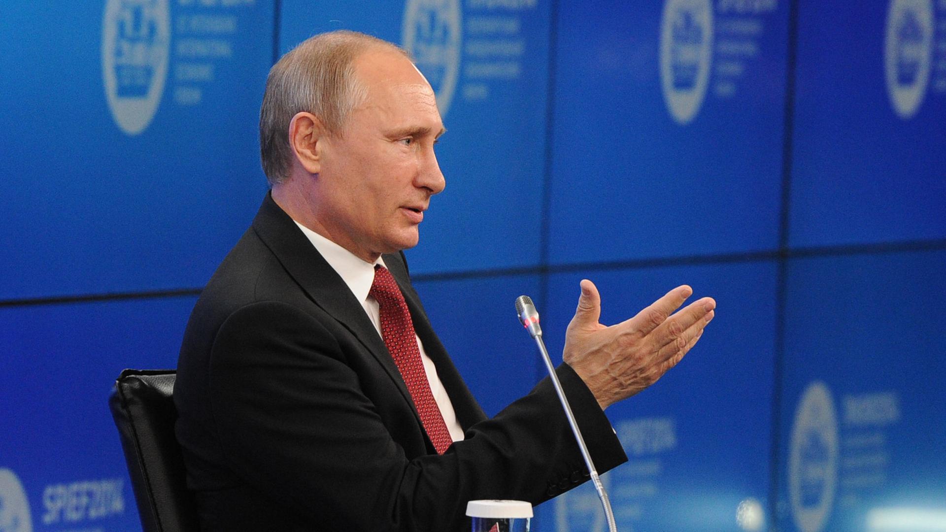 Wladimir Putin gestikuliert während seiner Rede an einem kleinen Tisch, neben dem er sitzt; hinter ihm eine blaue Wand mit Logos des Veranstalters