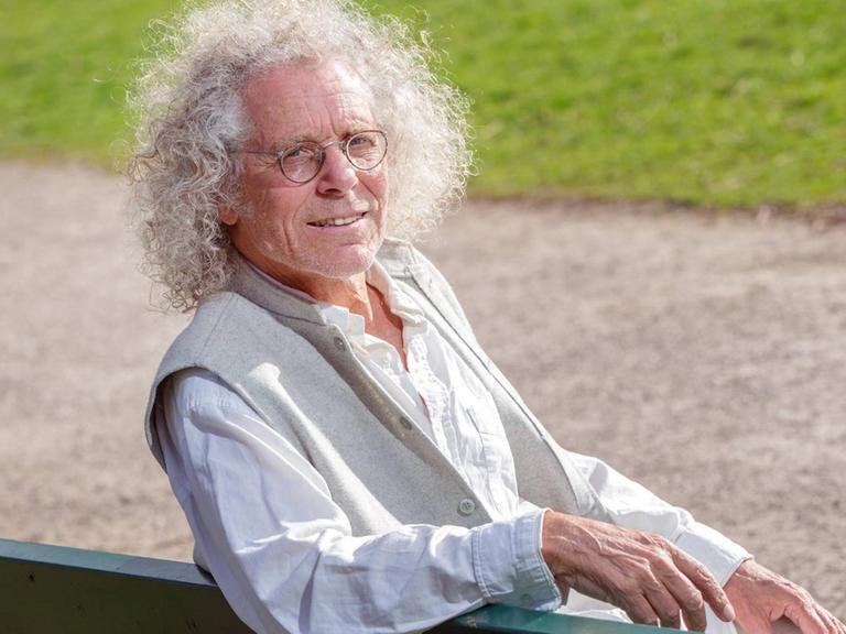 Rainer Langhans sitzt in München auf einer Parkbank und lächelt für eine Foto.