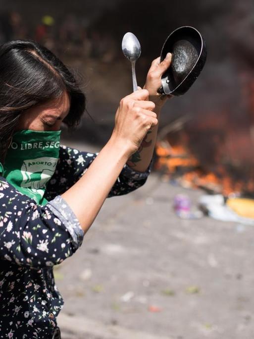 Gewaltsame Proteste in Santiago de Chile am 23. Oktober 2019. Eine Frau mit einem Tuch vor dem Gesicht schlägt mit einem Löffel auf einen Topf. Im Hintergrund brennen Barrikaden.