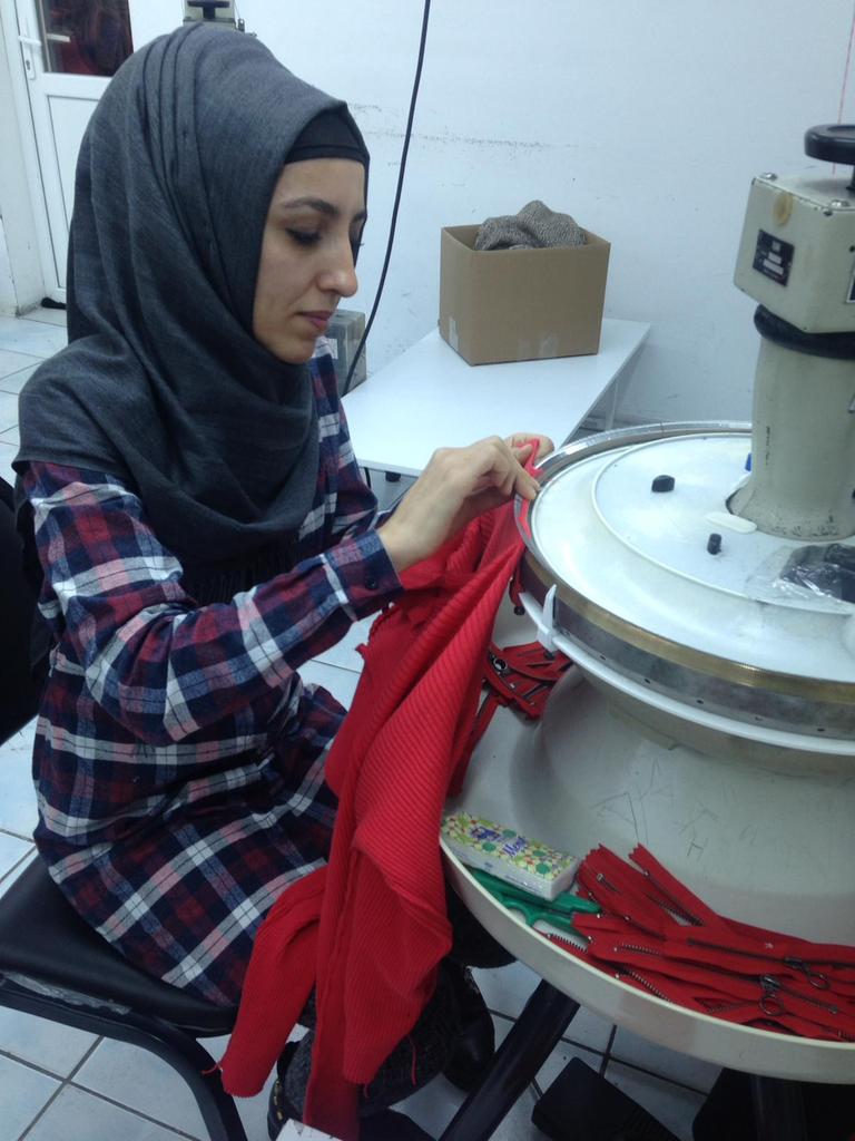 Eine junge Frau mit Kopftuche sitzt in einer Istanbuler Textilfirma an einer runden Strickmaschine und fertigt einen roten Pullover.