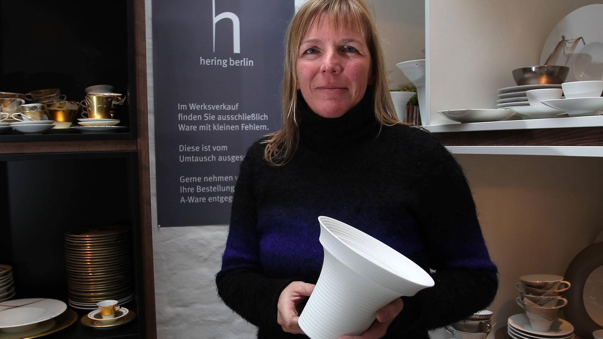 Die Töpferin, Porzellandesignerin und Tischdekorkünstlerin Stefanie Hering steht im Werksverkaufs ihres Geschirrs, hält eine Vase in der Hand. Um sie herum stehen in Regalen von ihr designte Stücke.