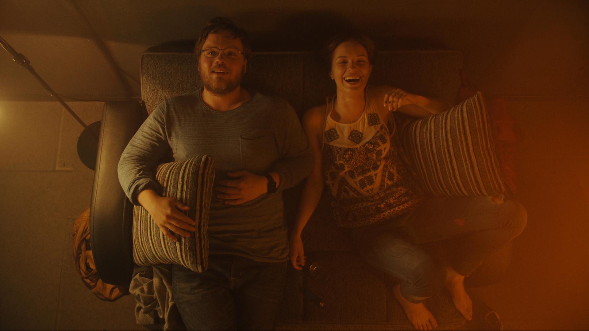 Adam Ild Rohweder und Paulina Galazka liegen in einer Szene aus "A Young Man with High Potential" auf einem Sofa und lachen. Sie werden von oben gezeigt, das Licht ist schummrig, aber warm.