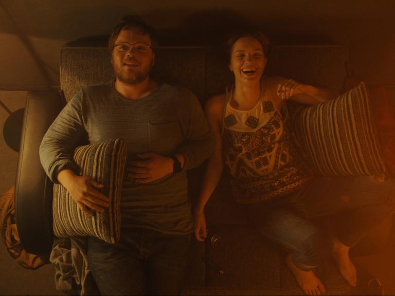 Adam Ild Rohweder und Paulina Galazka liegen in einer Szene aus "A Young Man with High Potential" auf einem Sofa und lachen. Sie werden von oben gezeigt, das Licht ist schummrig, aber warm.