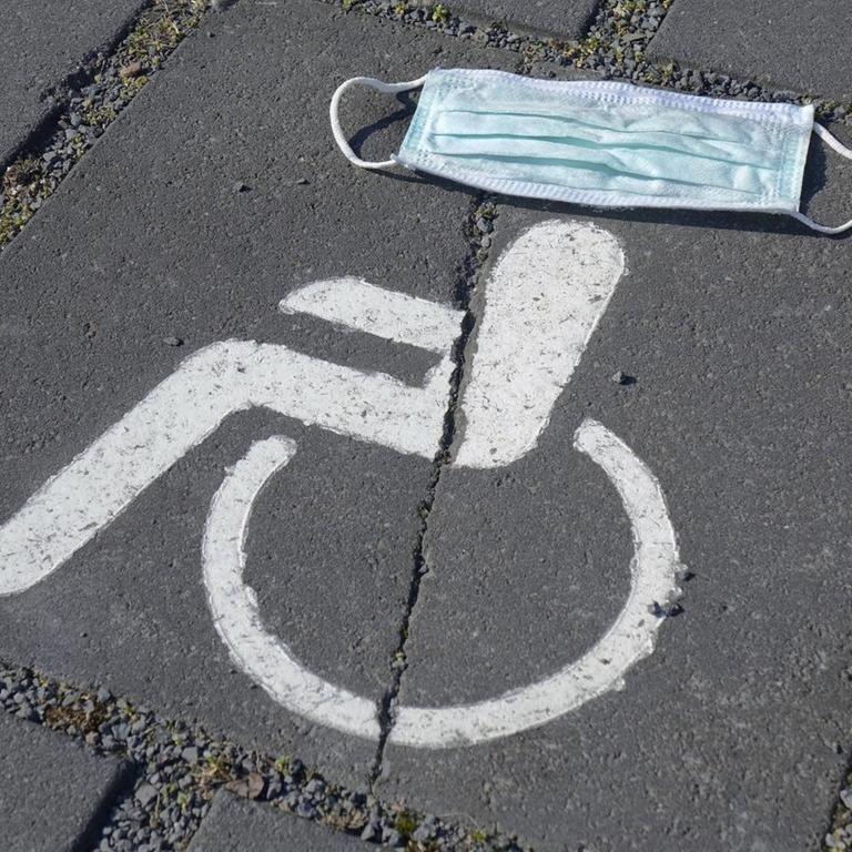 Symbol eines Behindertenparkplatzes, darüber ein Mundschutz