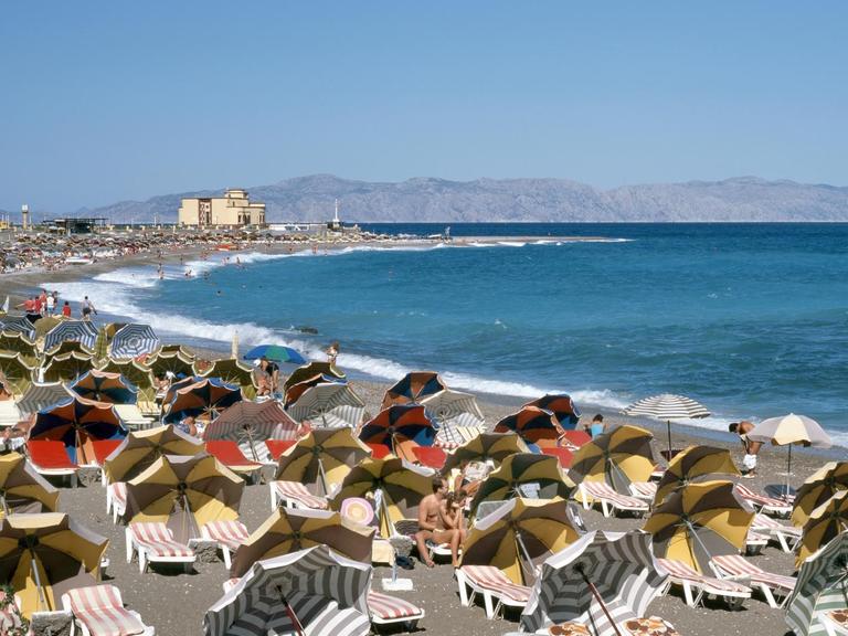 Touristen sitzen unter Sonnenschirmen und auf Liegen am Strand von Rhodos-Stadt.