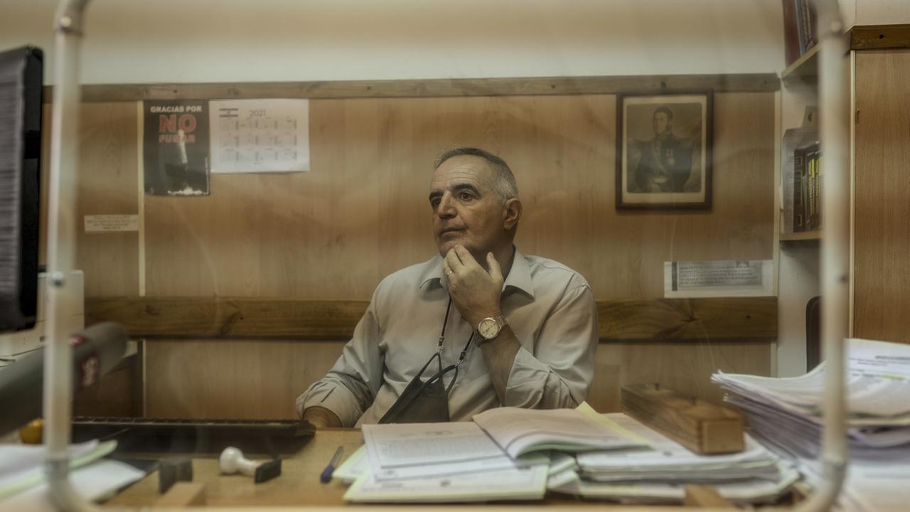 Ein Mann sitzt in einem holzgetäfelten Büro an seinem Schreibtisch und hat einen Berg von Unterlagen vor sich liegen.