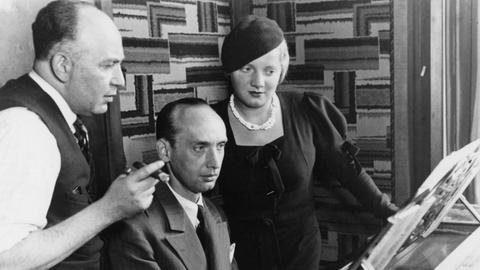 Ein Mann und eine Frau stehen neben einem am Klavier sitzenden Mann und schauen konzentriert auf die vor ihnen aufgestellten Notenblätter.