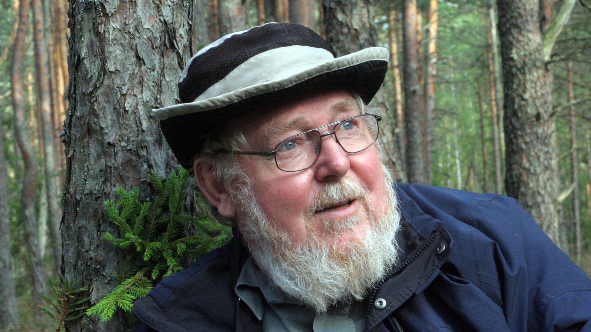 Der Biologe und Umweltschützer Michael Succow, Mütze auf dem Kopf, im Wald an einen Baum gelehnt.