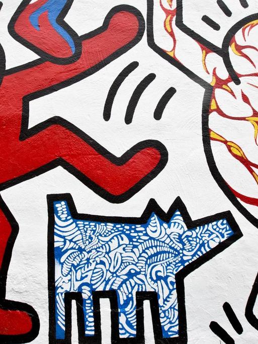 Das Bild zeigt einen Ausschnitt von Harings berühmten Strichmännchen als Wandmalerei. Links ein rotes und rechts ein weißes Strichmännchen mit rot-gelben Streifen. In der Mitte unten ist ein kleiner Hund mit blau-weißem Muster zu sehen.