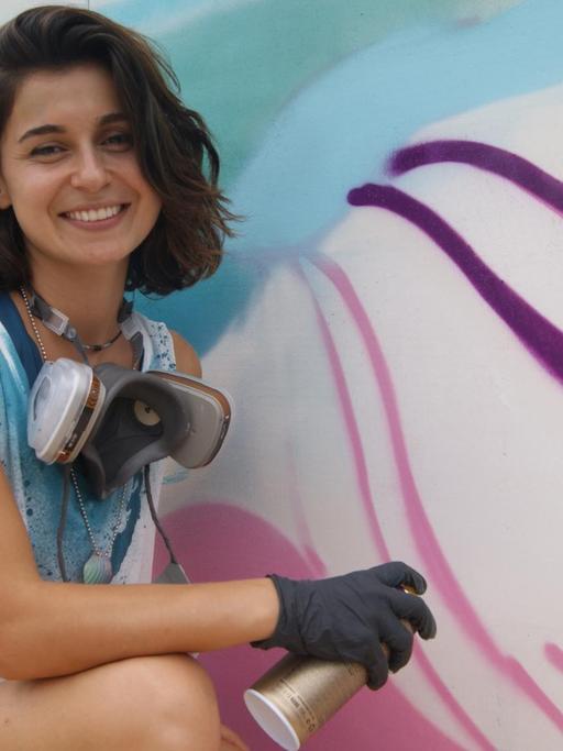 Auf dem Bild ist die Künstlerin Gamze Yalçın zu sehen. Sie lächelt in die Kamera und hält eine Sprühdose in der Hand. Rechts von ihr ist ein Graffiti zu sehen, an dem sie gerade arbeitet.