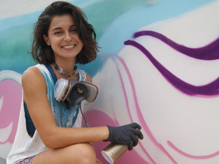 Auf dem Bild ist die Künstlerin Gamze Yalçın zu sehen. Sie lächelt in die Kamera und hält eine Sprühdose in der Hand. Rechts von ihr ist ein Graffiti zu sehen, an dem sie gerade arbeitet.
