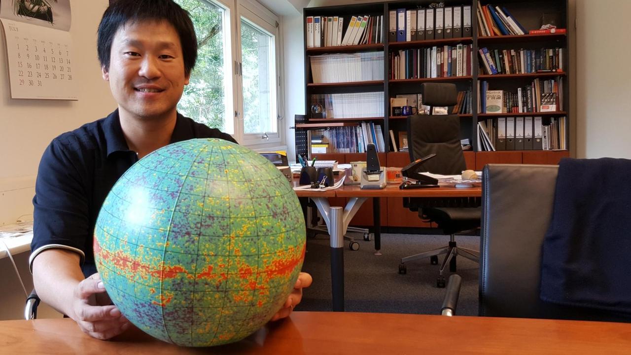 Eiichiro Komatsu sitzt an einem Schreibtisch und hält einen Globus in den Händen