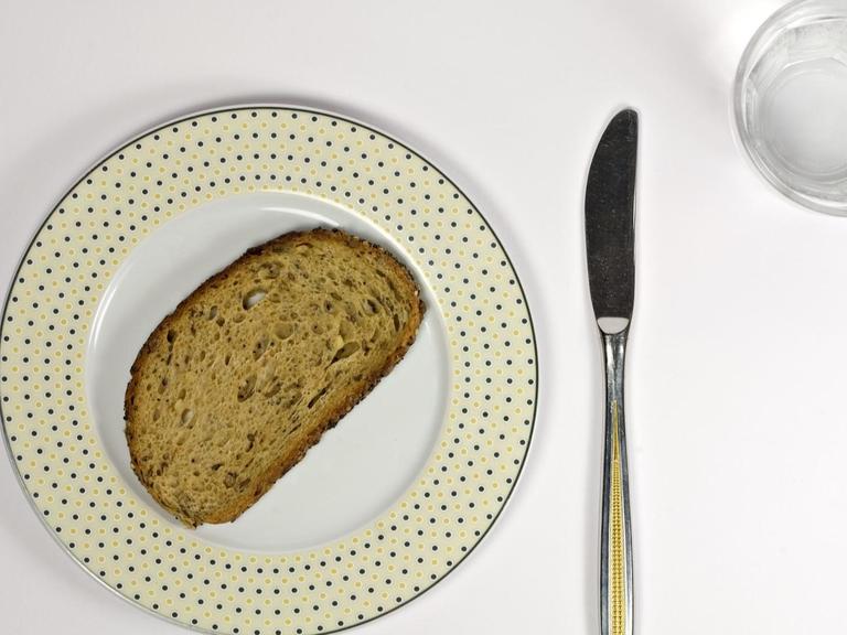 Neben einem Teller, auf dem eine Scheibe Brot liegt, liegen Messer und Gabel. Zudem steht ein mit Wasser gefülltes Glas dabeil