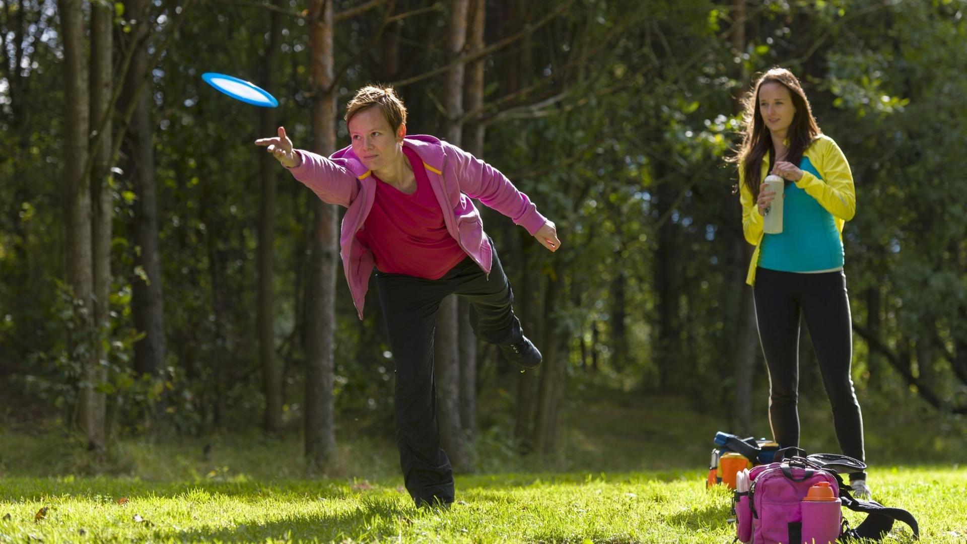 Zwei junge Frauen spielen auf einer Wiese am Waldrand Frisbee.