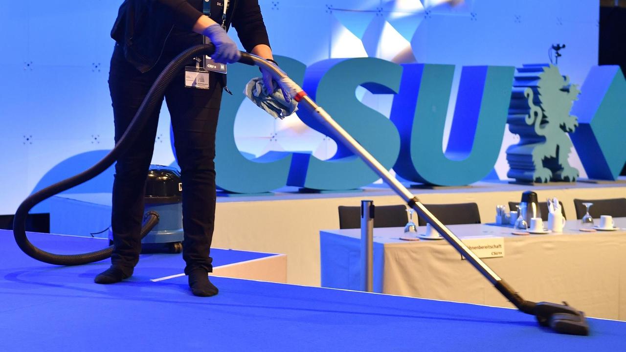 Eine Reinigungskraft beim Staubsaugen des blauen Teppichbodens vor dem CSU Logo. 
