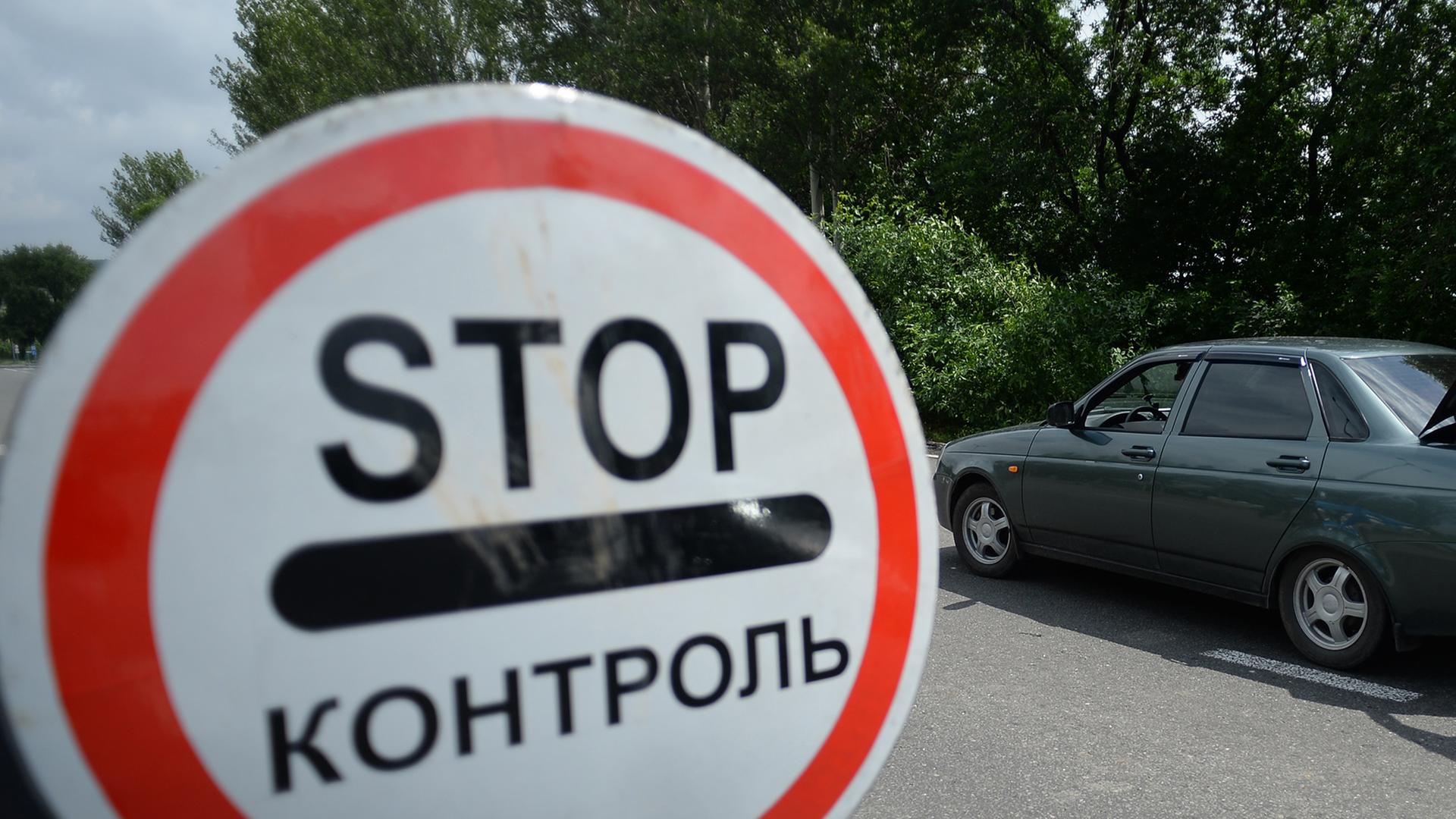 Links im Vordergrund ein rundes weißes Schild mit rotem Rand und der Aufschrift "Stop", im Hintergrund rechts ein Auto