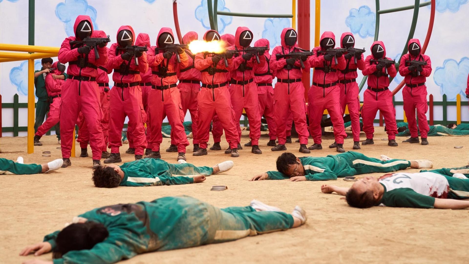 Eine Szenenfoto aus einem Film: Männer in roten Uniformen tragen Maske und Gewehre. Vor ihnen liegen Menschen in grünen Overalls im Sand.