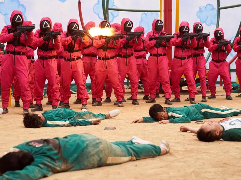 Eine Szenenfoto aus einem Film: Männer in roten Uniformen tragen Maske und Gewehre. Vor ihnen liegen Menschen in grünen Overalls im Sand.