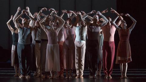Mehrere Tänzer stehen als Gruppe in einer aufrechten Pose mit erhobenen Armen auf der Bühne.