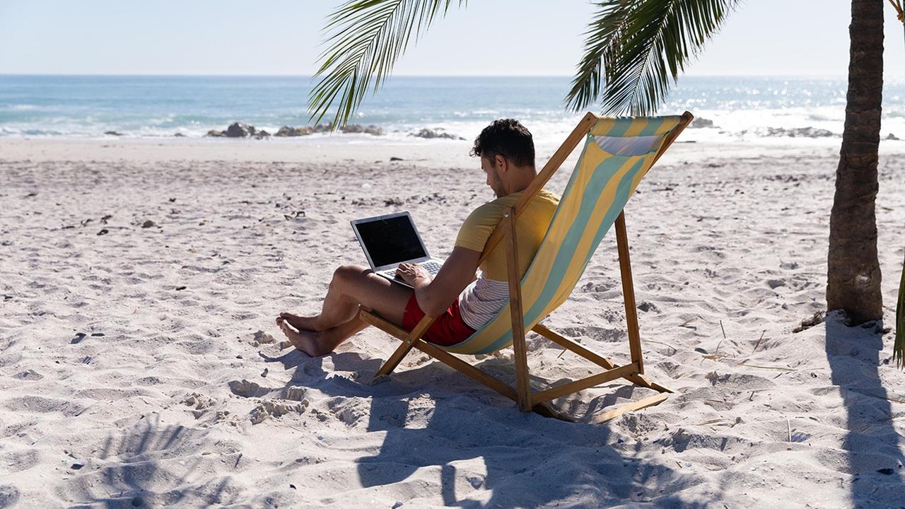 Ein Mann sitzt unter einer Palme an einem Laptop am Strand.