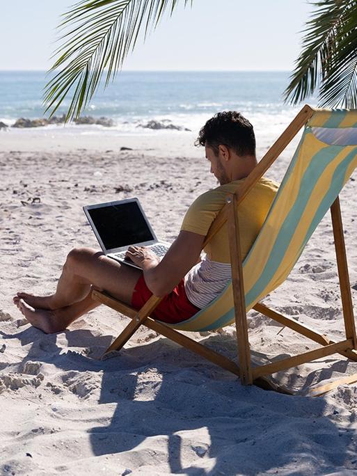 Ein Mann sitzt unter einer Palme an einem Laptop am Strand.