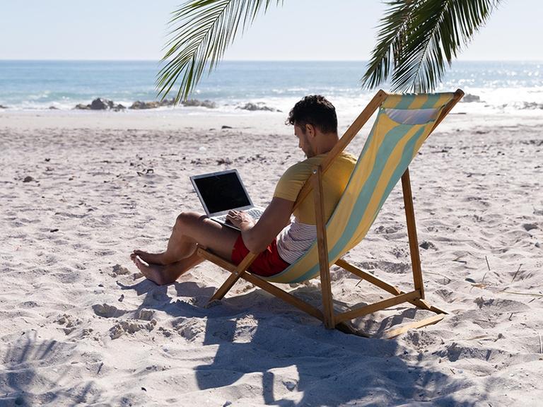 Ein Mann sitzt unter einer Palme an einem Laptop am Strand.