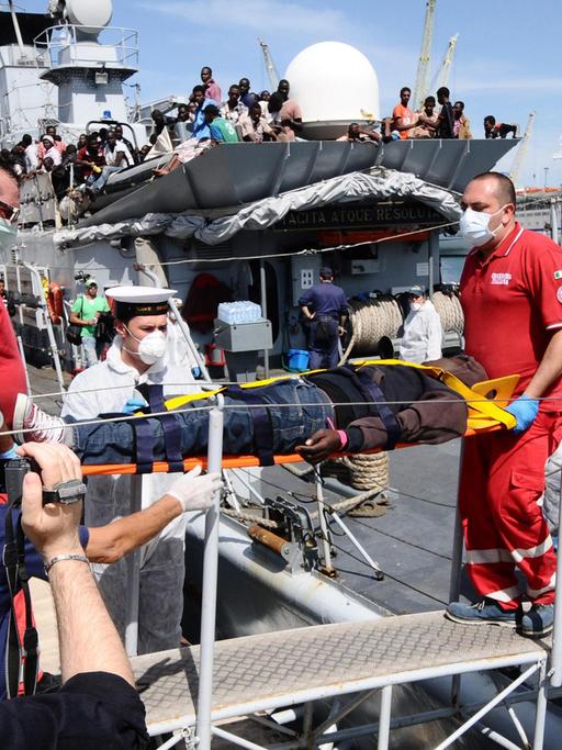 Sanitäter in roten Uniformen und mit Mundschutz tragen einen auf einer Trage liegenden Flüchtling von Bord eines Schiffs.