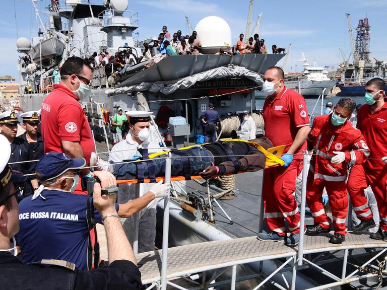Sanitäter in roten Uniformen und mit Mundschutz tragen einen auf einer Trage liegenden Flüchtling von Bord eines Schiffs.