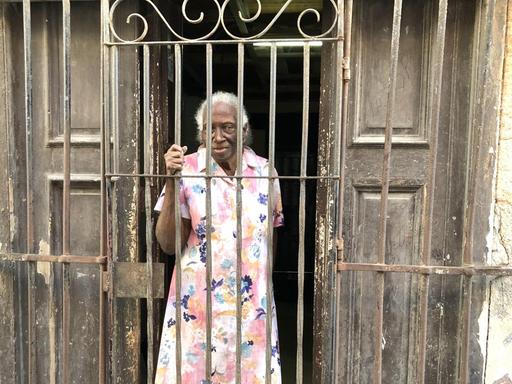 Eine ältere Kubanerin steht in der Altstadt von Havanna vor ihrer etwas baufälligen Haustür und einer Hauswand, von der der Putz bröckelt, und greift mit der rechten Hand an das hohe Gitter, das sie von der Straße trennt.