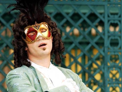 Ein Sänger mit Perücke, Maske und Rokoko-Mantel steht im Freien und singt.