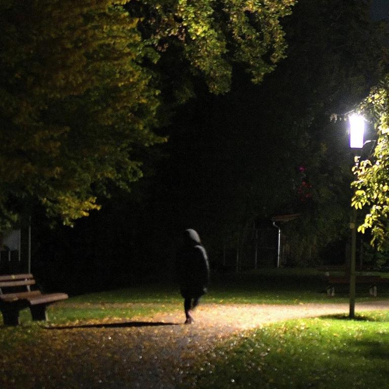 Eine Frau läuft nachts alleine an einem beleuchteten Weg in die dunkle Nacht.