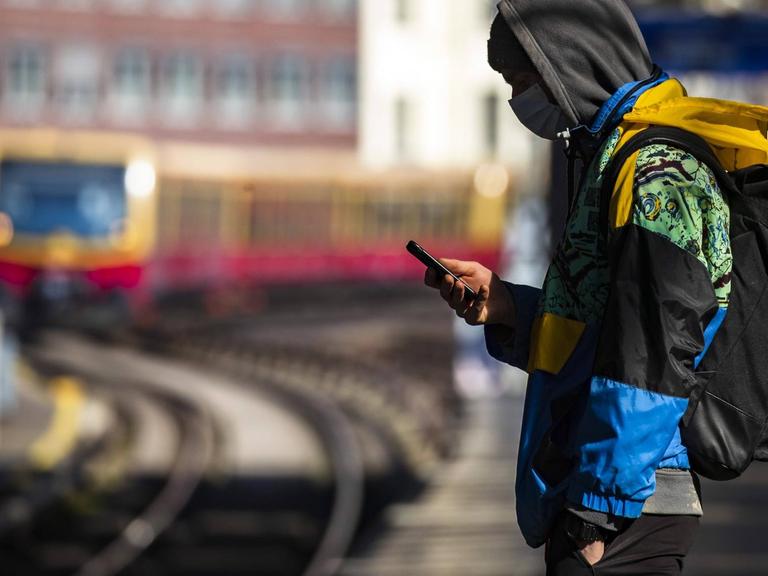 Ein Jugendlicher steht mit Mundschutzmaske auf einem S-Bahnsteig in Berlin und blickt auf sein Handy.