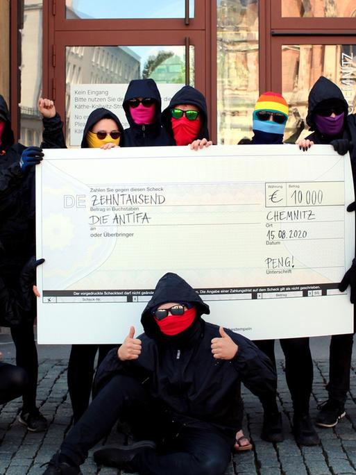 Mehrere maskierte Menschen stehen mit einem symbolischen Scheck vor einem Gebäude.