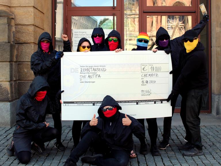 Mehrere maskierte Menschen stehen mit einem symbolischen Scheck vor einem Gebäude.