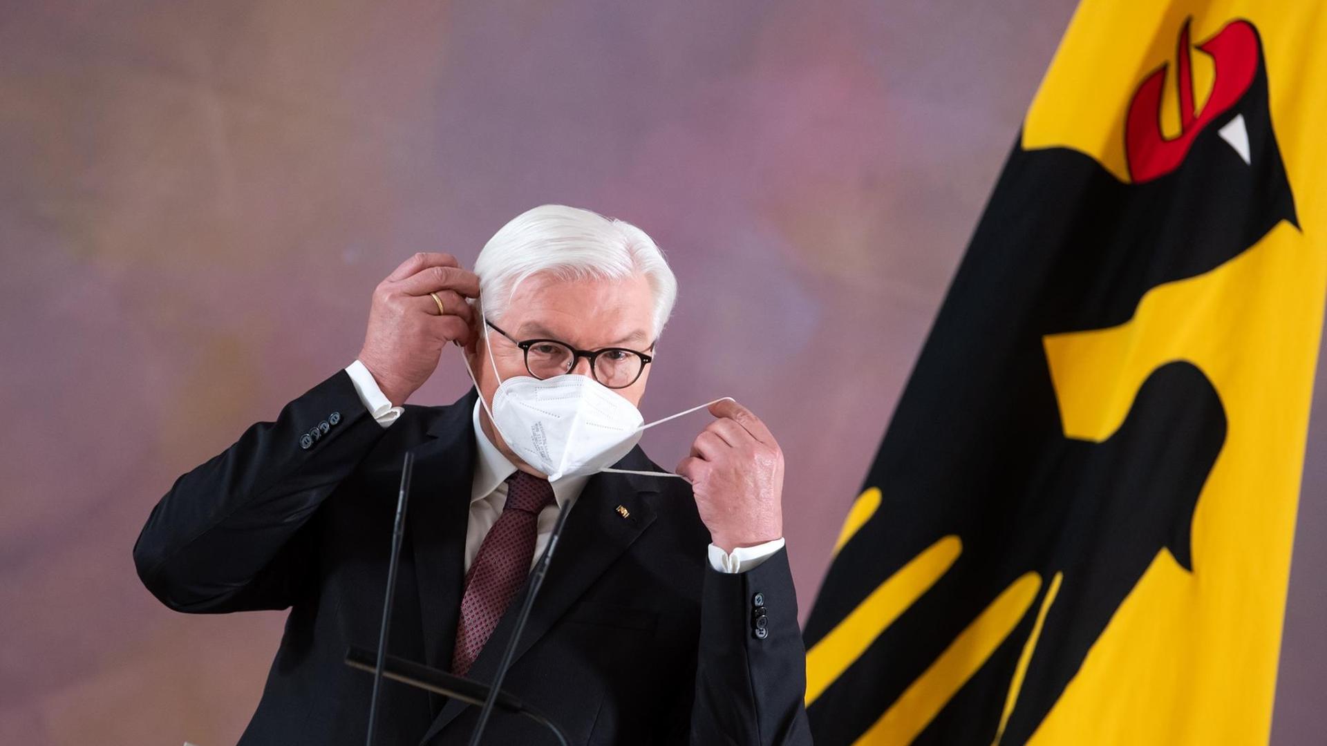 Bundespräsident Frank-Walter Steinmeier setzt sich im Schloss Bellevue nach seinem Statement, dass er für eine zweite Amtszeit bereit steht, eine FFP2-Maske auf. Am rechten Bildrand ist eine gelbe Flagge mit einem schwarzen Adler zu sehen.