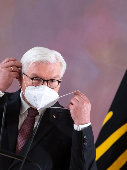 Bundespräsident Frank-Walter Steinmeier setzt sich im Schloss Bellevue nach seinem Statement, dass er für eine zweite Amtszeit bereit steht, eine FFP2-Maske auf. Am rechten Bildrand ist eine gelbe Flagge mit einem schwarzen Adler zu sehen.