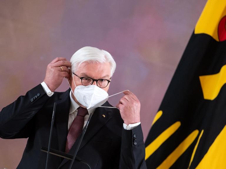 Bundespräsident Frank-Walter Steinmeier setzt sich im Schloss Bellevue nach seinem Statement, dass er für eine zweite Amtszeit bereit steht, eine FFP2-Maske auf. Am rechten Bildrand ist eine gelbe Flagge mit einem schwarzen Adler zu sehen.