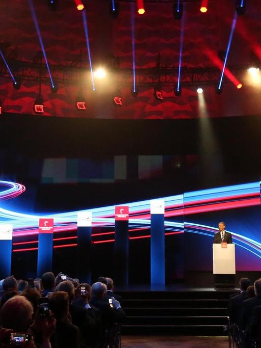 Sie sehen Barack Obama auf einer großen Bühne am Rednerpult, dahinter bunte Licht-Kurven als Beleuchtung.