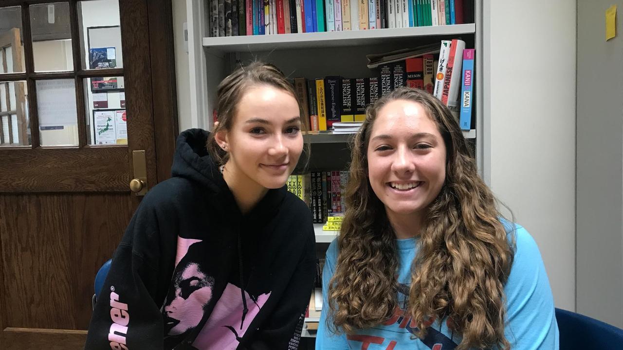 Zwei junge Studentinnen sitzen an einem  Tisch vor einem Bücherregal.