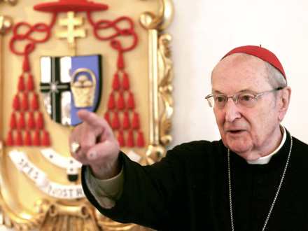 Joachim Kardinal Meisner im Ornat steht vor seinem Wappen und zeigt mit dem rechten Finger an der Kamera vorbei.