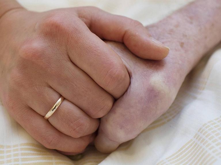 Zwei Hände halten sich umschlossen auf einem Laken. Die Linke Hand trägt einen Ehering.