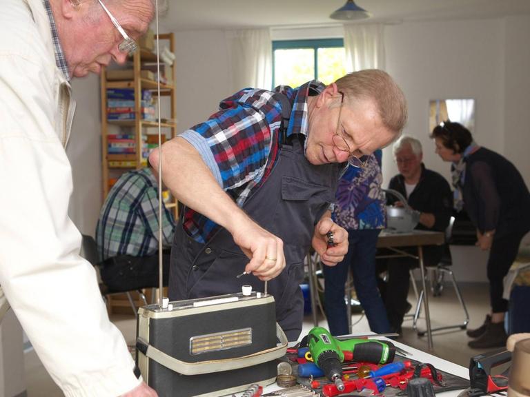 Ein Mann repariert ein altes Radio bei einem Repair Café Treffen. Repair Cafés sind ehrenamtliche Treffen, bei denen die Teilnehmer alleine oder gemeinsam mit anderen ihre kaputten Dinge reparieren.