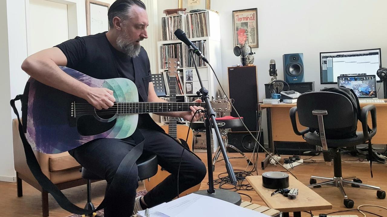 Der Musiker Fink, bürgerlicher Name Finian Paul Greenall, in seinem Studio in Berlin. Er spielt im Sitzen Gitarre, vor ihm ein Mikroständer. Im Hintergrund Gitarren, ein Bücherregal, Lautsprecherboxen und ein Computer aauf dem Schreibtisch. 