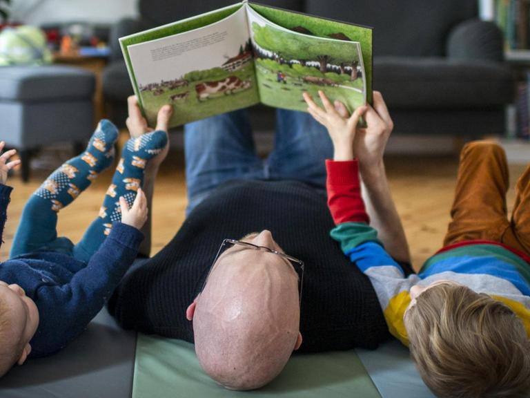 Ein Vater liest mit seinen beiden Kindern auf dem Boden liegend ein Buch.