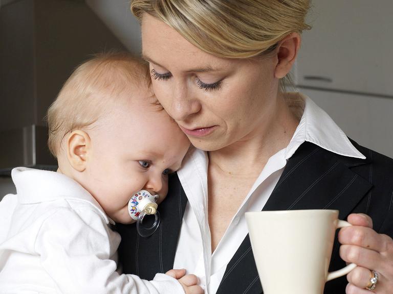 Eine Mutter im Business-Anzug mit ihrem Baby auf dem Arm, in der Hand eine Kaffeetasse.